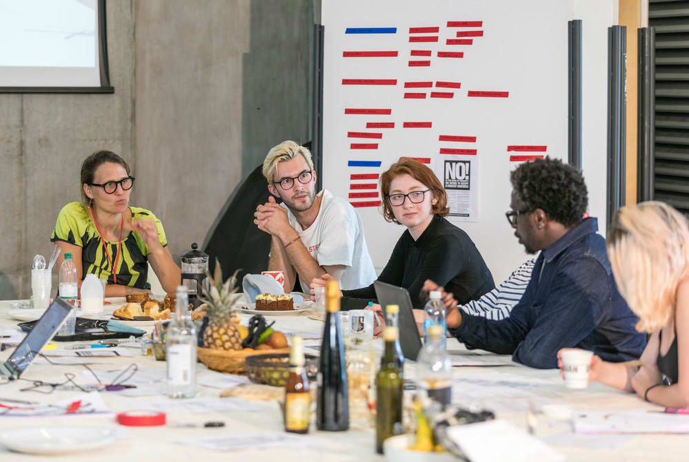 5 people (mixed gender, race) talking at a table with papers, laptops, fruit and drinks in front of them