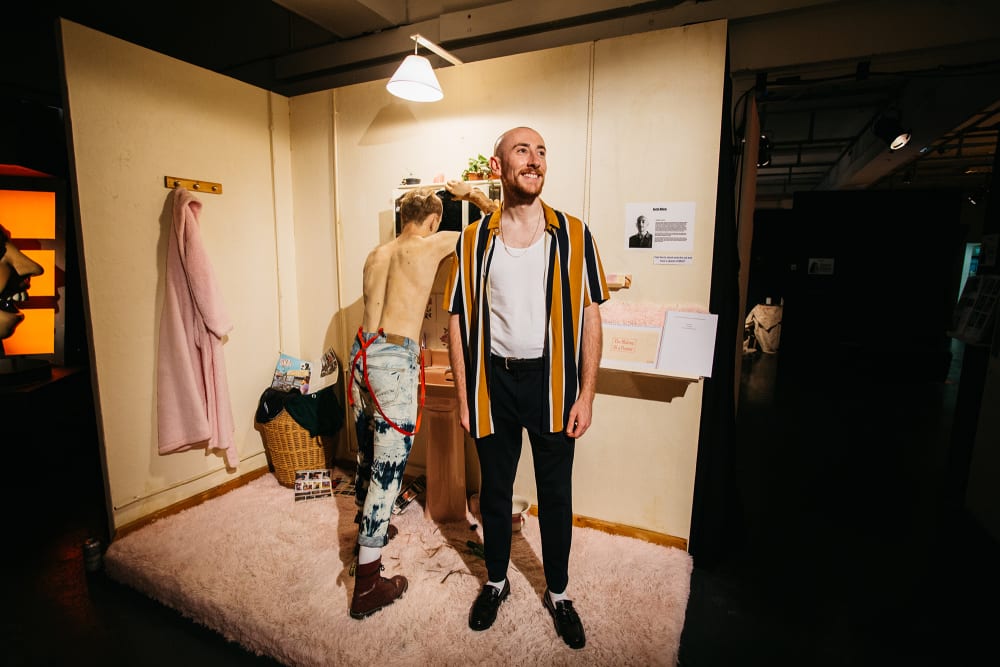 A recreation of a 1980s bathroom suite in which a realistic model of a young man stands at the sink shaving his head. The model is topless and wearing bleached denim jeans with red braces. In the foreground stands the student who made this scene - Jack Allen. He is smiling and wearing black trousers and an orange and black striped shirt open over a white vest.
