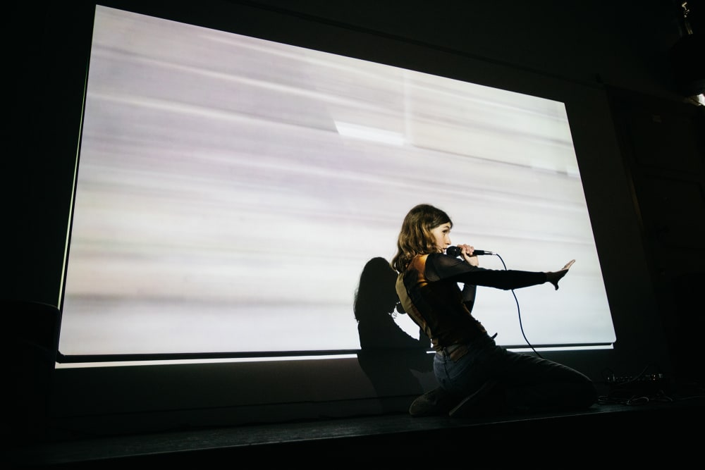 A woman dressed in black holds a microphone with her right hand outstretch kneels in front of a large screen showing a blurred projection of white light.