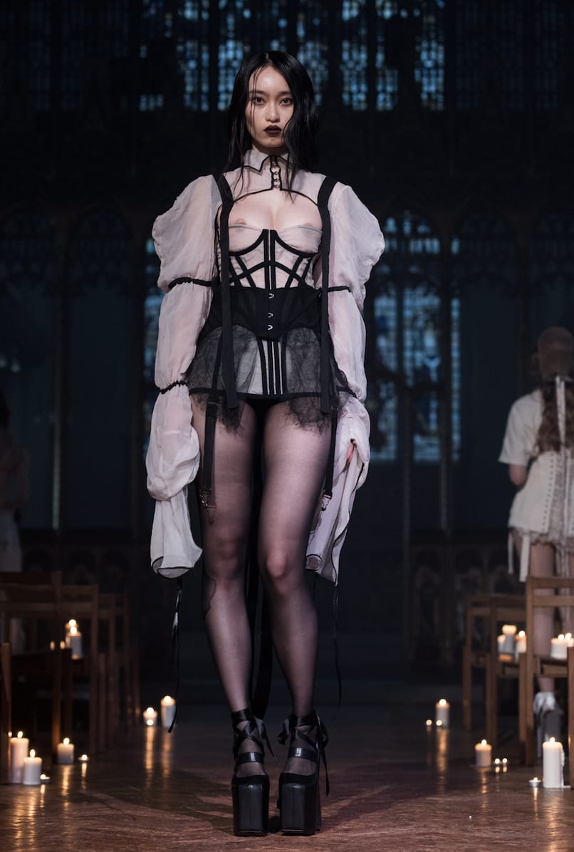 Model on catwalk wearing a black corset over a sheer blouse