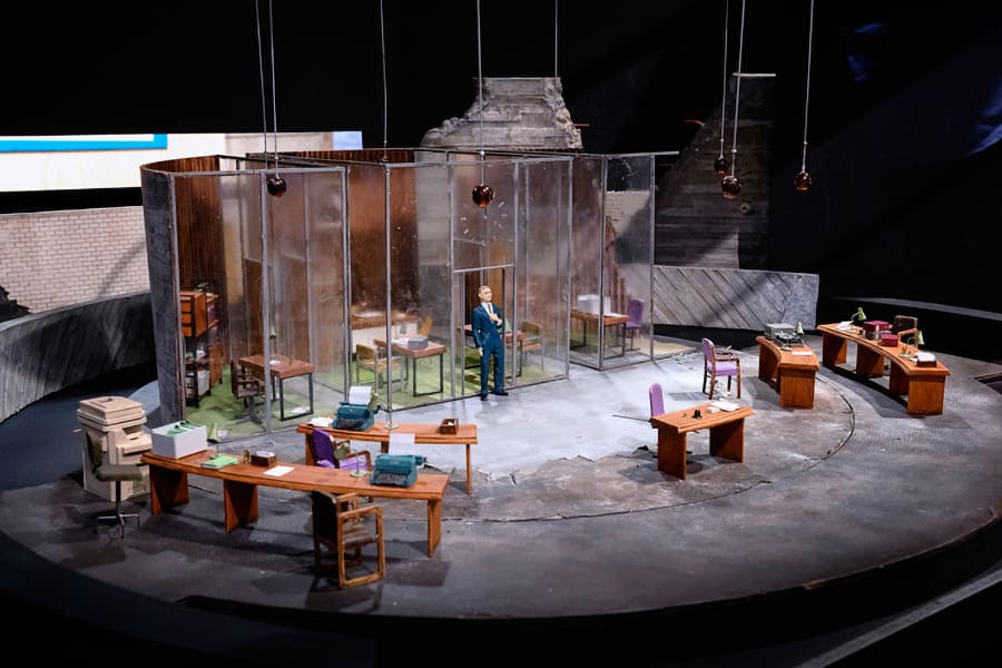 A small scale set design of a man in an office surrounded by desks.