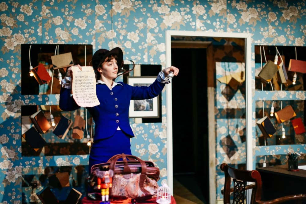 A woman in a blue suit and hat holding up a piece of paper with blue flowery wallpaper behind her.
