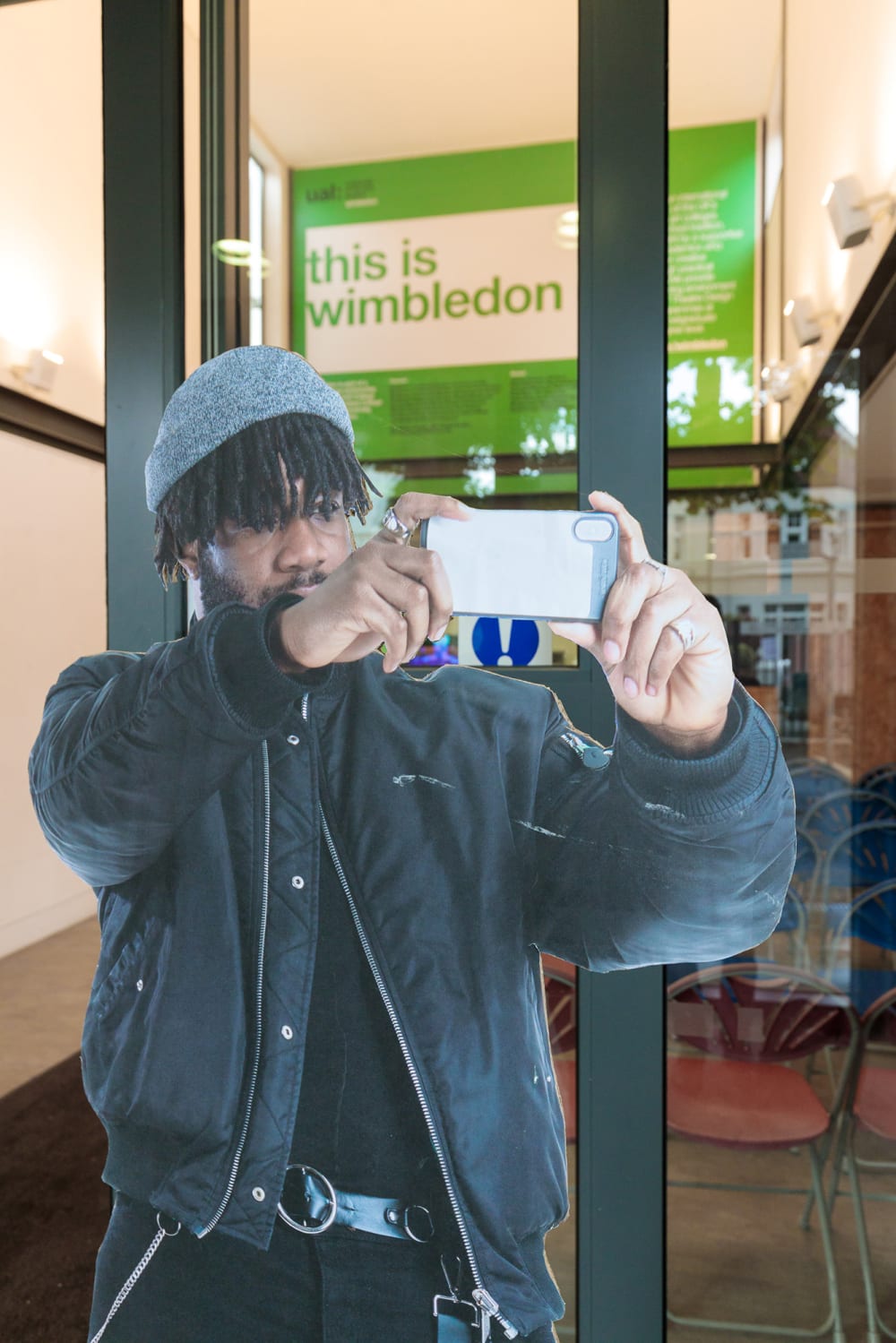 A cardboard cutout of a man dressed all in black with a grey had and short black dreadlocks holding up his phone as though to take a landscape photo. Visible in the background in a sign in green which says in large letters 'this is Wimbledon'.