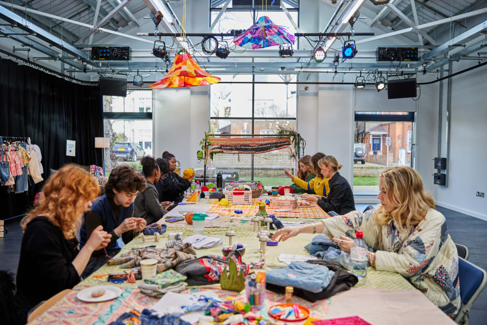 Image shows 9 people sitting round a large rectangle table. On the table, there are various craft items and the people sitting round the table are engaging with crafts and making. In the background, there are ceiling to floor windows with lots of light pouring into the room. Overhead, studio lighting can be seen.