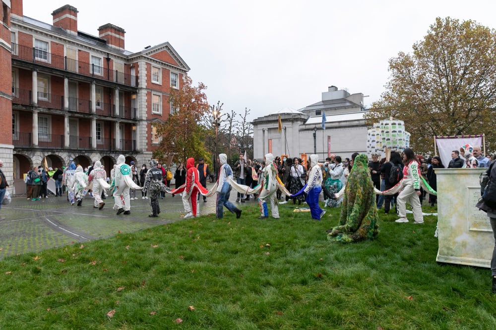 A line of people wearing boilersuits which are all joined together parade in a line across a large open-air space with some grass areas and some paved areas. One of the college buildings - large, red brick and old - is in the background.