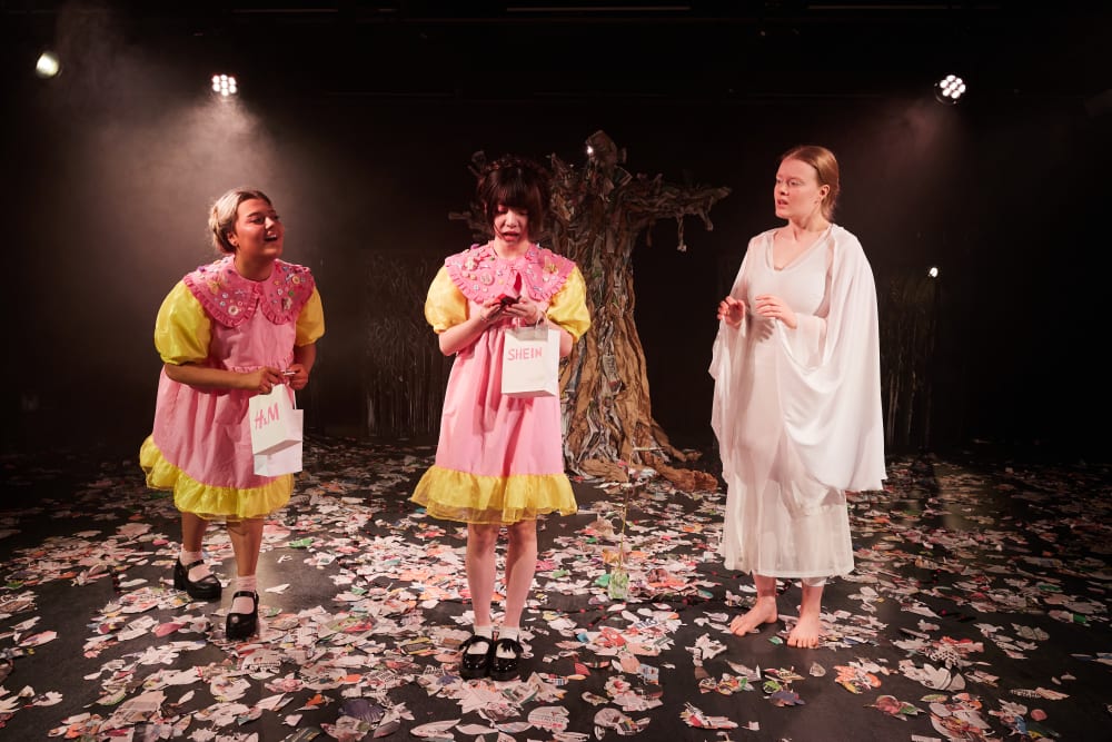 Image shows 3 performers standing on a stage. 2 of the performers are wearing pink and yellow dresses and carrying shopping bags, while the other performers is wearing a long white dress. In the background, you can see a tree. 