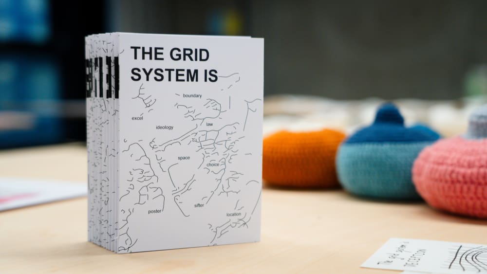 Image shows a white book entitled ‘The Grid System is’. The book is sitting on top of a table with colourful items blurred in the background.