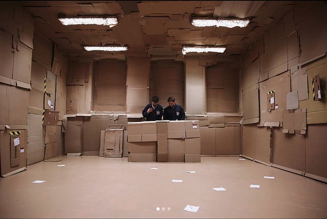 A room covered in cardboard boxes with two men at the back behind a stack of them.