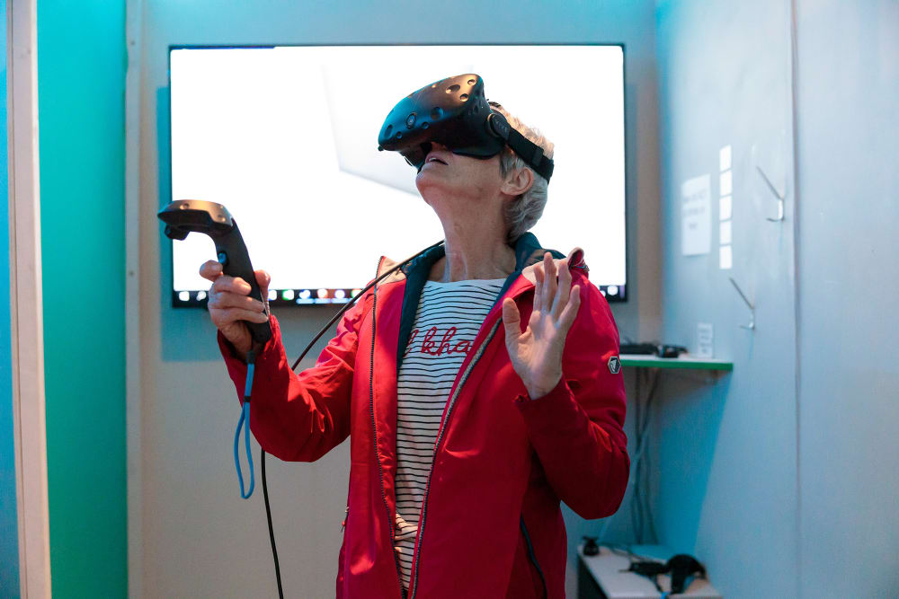 A person in a red jacket wears a virtual reality headset and holds a controller. They are looking upwards with their hands held up as if they are exploring a space that we can't see.
