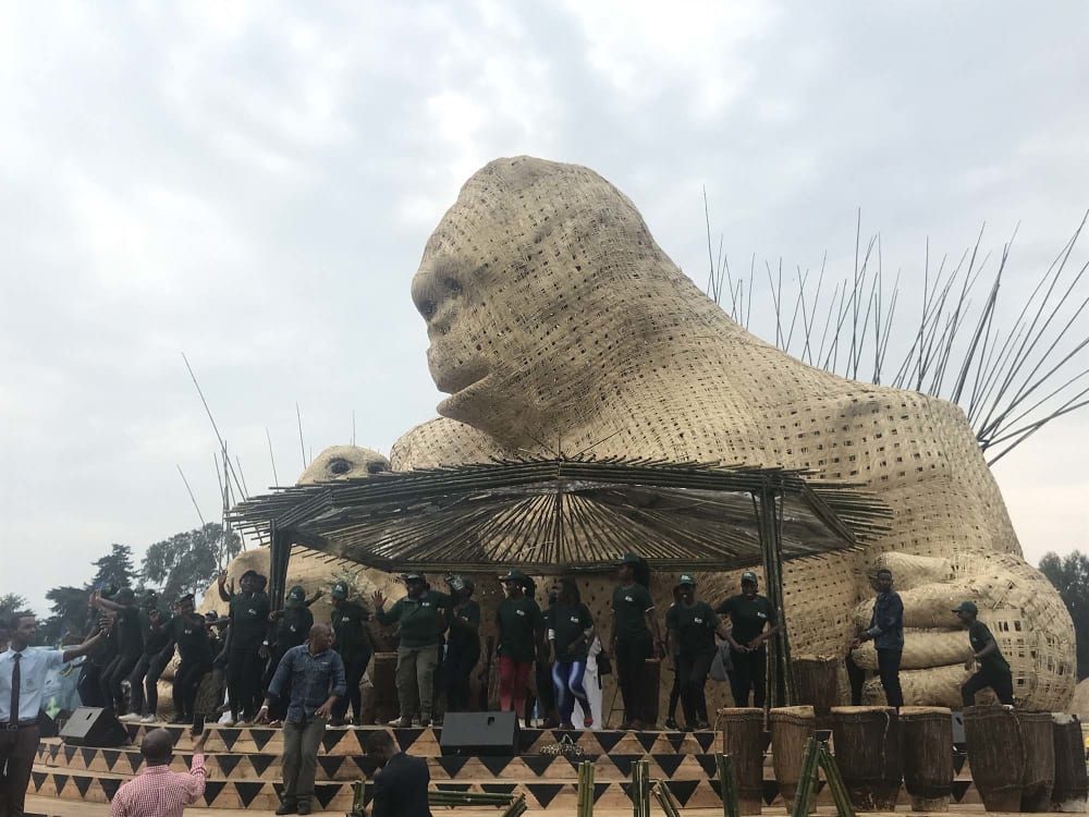 A group of people on a stage under a bamboo shelter. Behind them is a large woven sculpture of a gorilla mother and child.