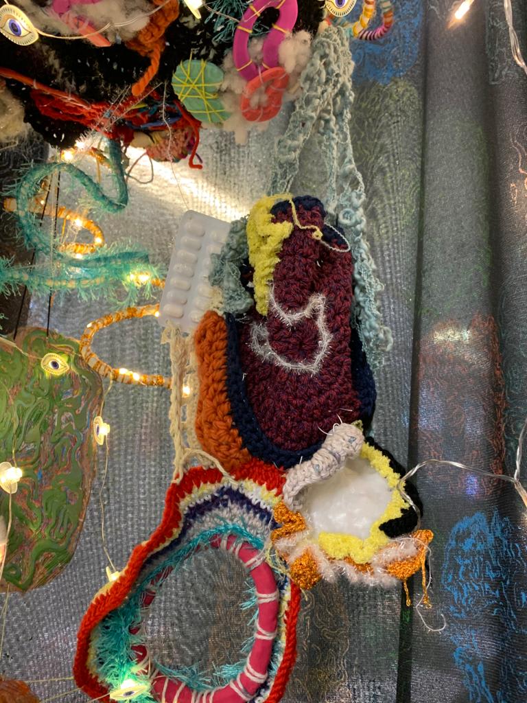Draped black curtains with different coloured brain scans hangs with several knitted headpieces inspired by migraines. A pack of pills also hangs within the piece along with fairy lights that look and represent eyes.