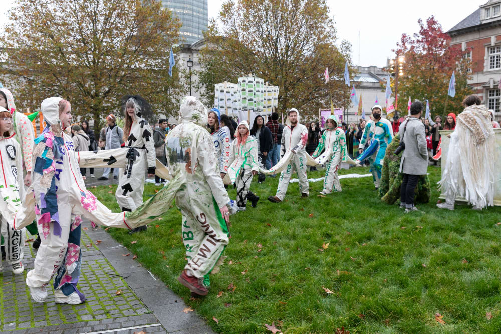 A line of people wearing boilersuits which are all joined together parade in a winding chainacross a large open-air space with a grass area and a paved area.