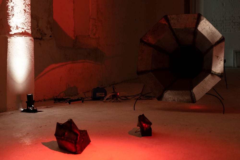A dark room lit in red light showing a large gramophone-like sculpture with other objects on the floor around it.