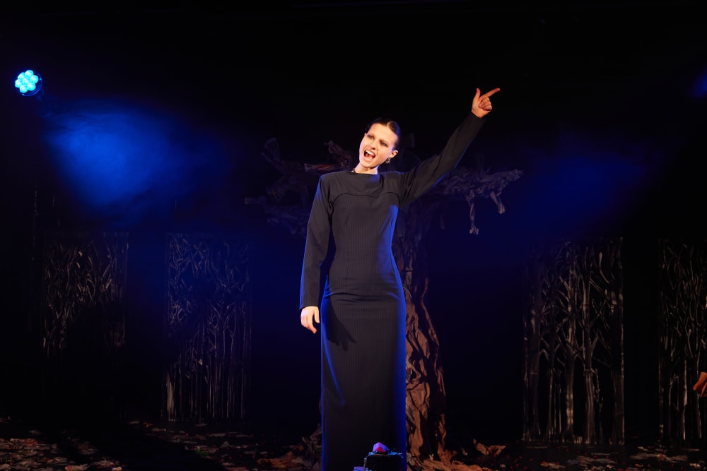 Image shows a person standing in a long black dress with pinned back hair pointing a finger up. They are standing in front of a tree and have an intense facial expression.