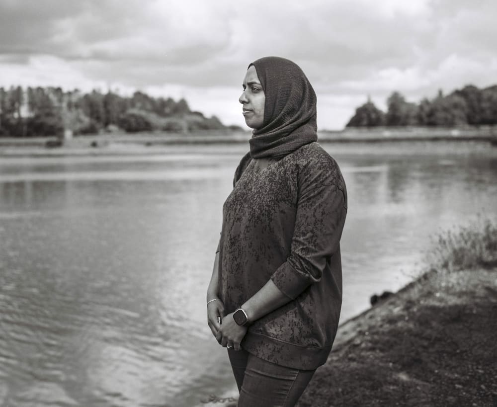 A black and white photo of a woman in a headscarf standing in front of a lake, looking out across it. Her hands are folded in front of her