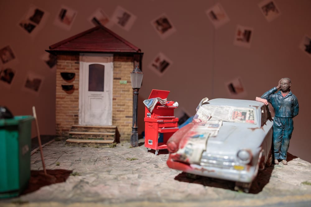 Close up photo of a model showing a man leaning with his left elbow on a car which is being repainted and is partically covered in newspapers. To the left of this is a red workstation on wheels, and beside that a small brick house with a white front door. On the far left of the image in the foreground and out of focus is a green wheelie bin.