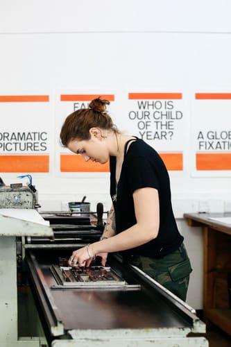 Student working in the LCC Letterpress Studio, backdrop has orange posters. 