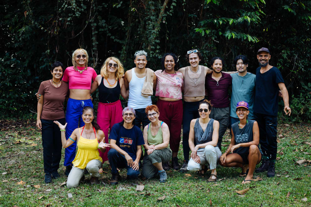 Fourteen people pose for a photograph smiling at the camera with trees in the background. 