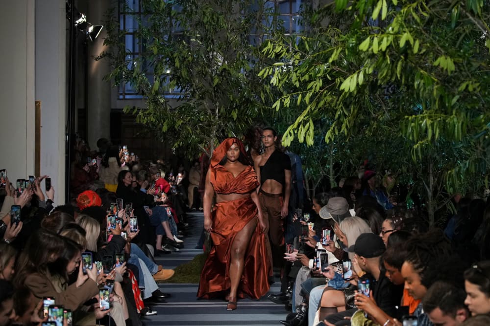 Image from a catwalk show with models walking down the centre of the catwalk surrounded by plants and trees. 
