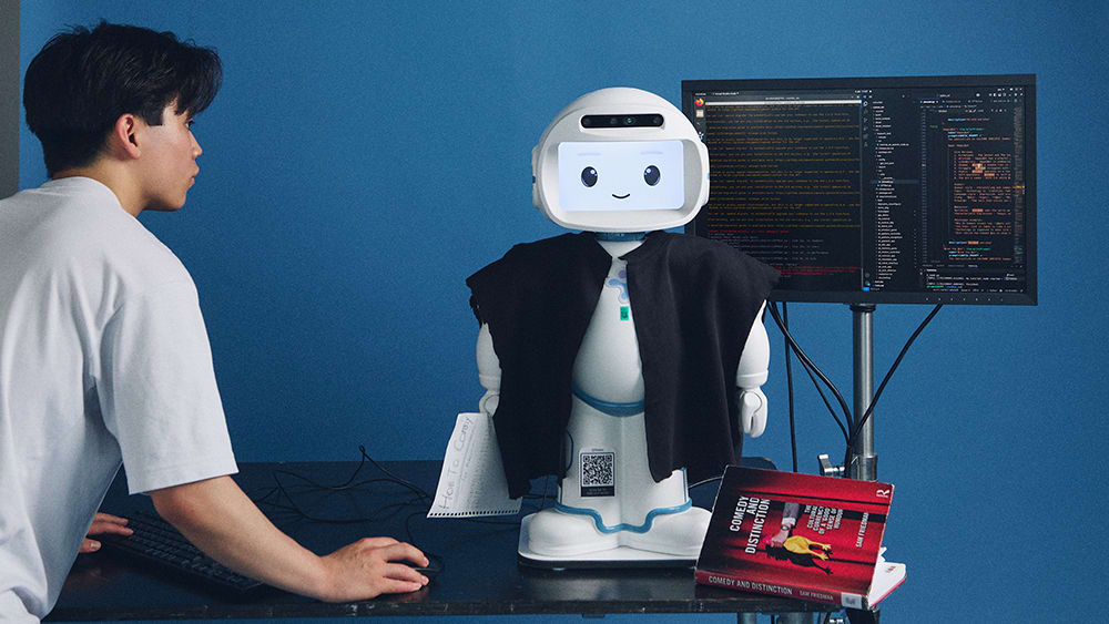 CCI student Michael Wong Kam interacts with a small robot wearing a black jacket. The robot is positioned on a desk with a monitor showing code and a book on comedy.