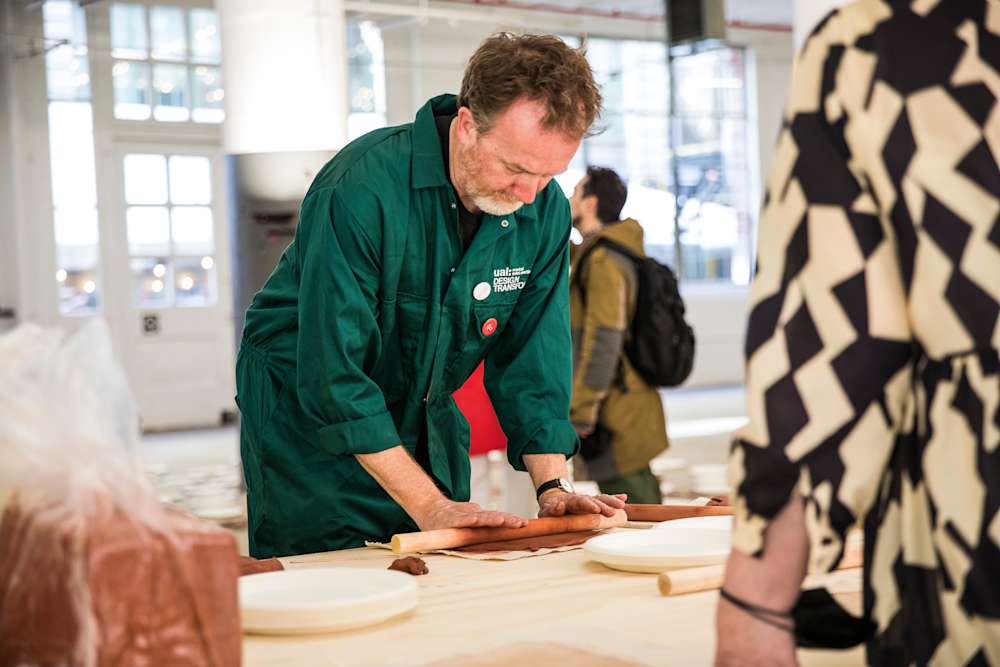 Tony Quinn is wearing a green boilersuit and rolling out clay on a table, with other people around him.