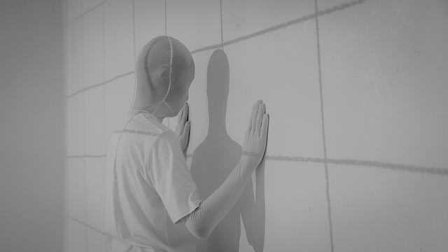 Figure all in white, wearing a white mask with their hands paled against the wall.