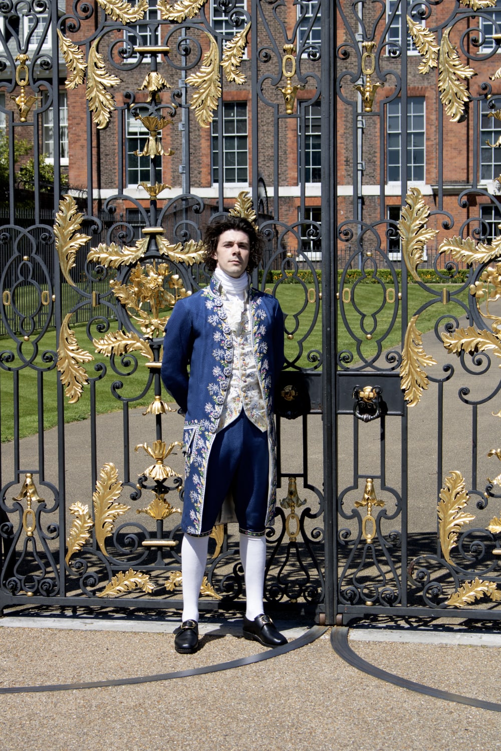 A man standing wearing a blue embroidered silk suit with white silk stockings and black shoes, in front of ornate black and gold gates.