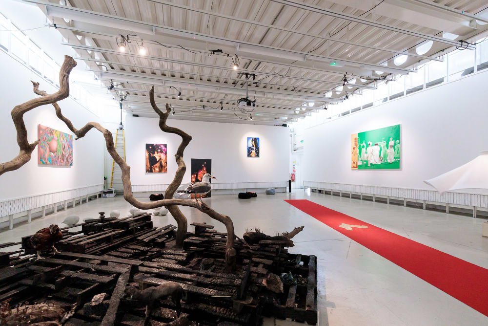 A large room with artwork installed throughout. In the foreground, a large pile of charred wood from which some bare branches protrude. To the right, a long strip of red carpet runs to the back of the room. On the far right wall a green painting hangs.