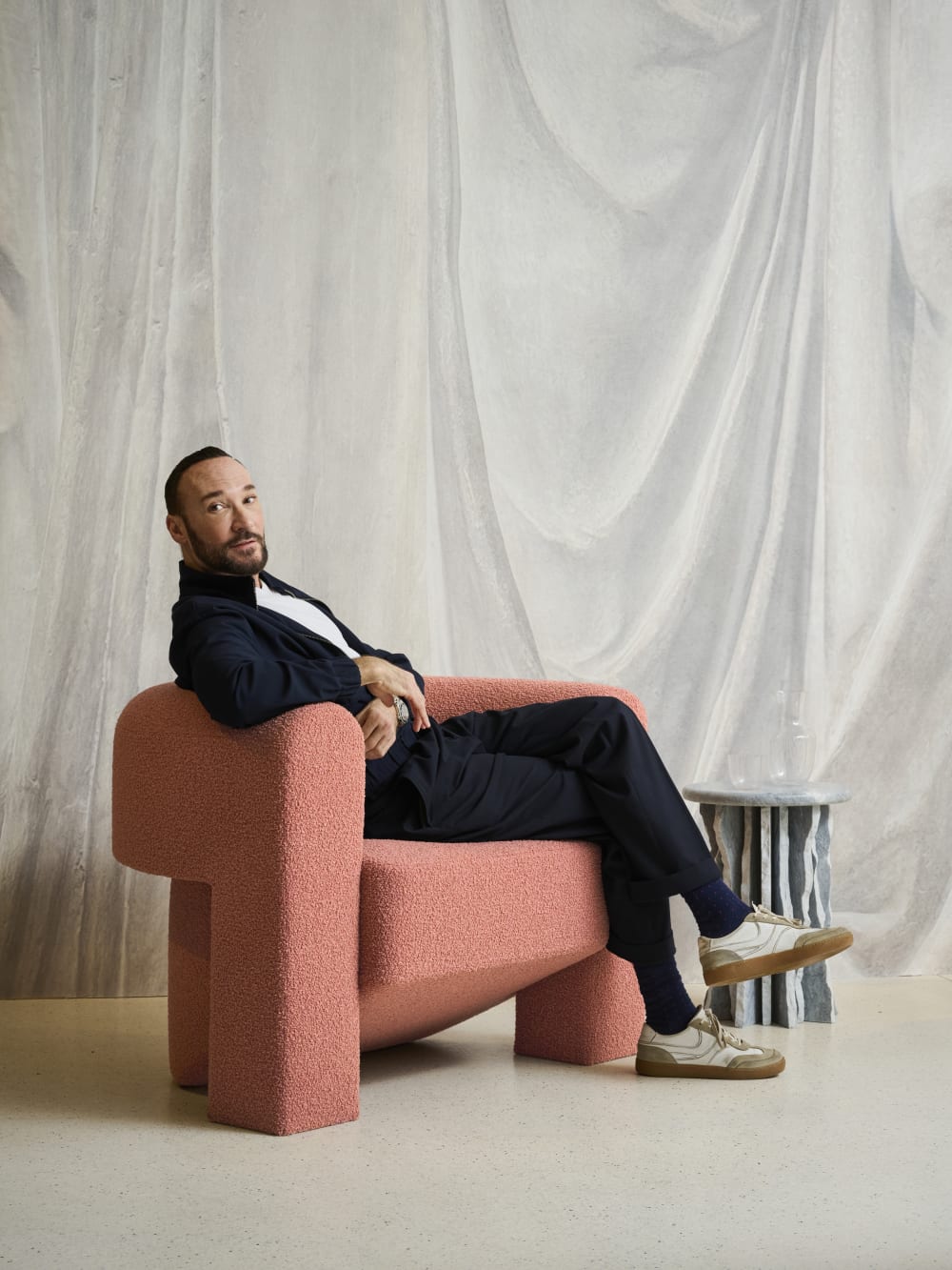Bodo sat in pale pink chair looking over his shoulder. White fabric is hanging from the ceiling behind him