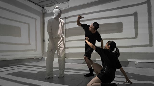 A figure all in white with their face covered standing above two figures wearing black.
