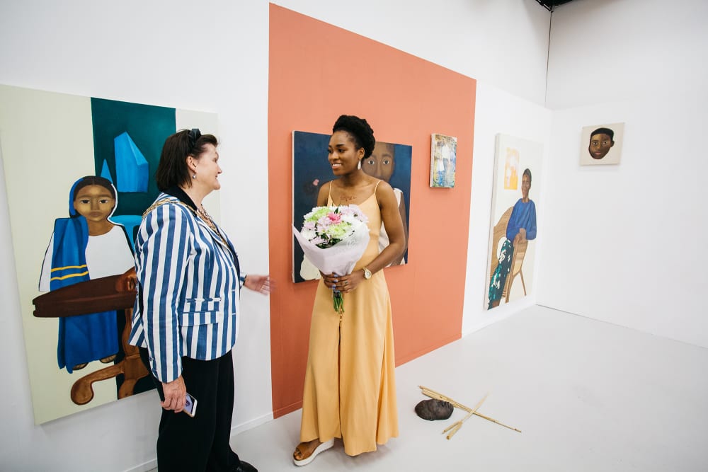 Two women stand in front of paintings. The woman on the left wears a blue and white striped jacket and black trousers. The woman on the right is in a peach, floor-length dress and holds a bunch of flowers. There are five paintings behind them: three are on a white wall and the wall behind the middle two has been painted a terracotta colour. On the floor in front of these is a bronze sculpture of a face and two dried reeds.