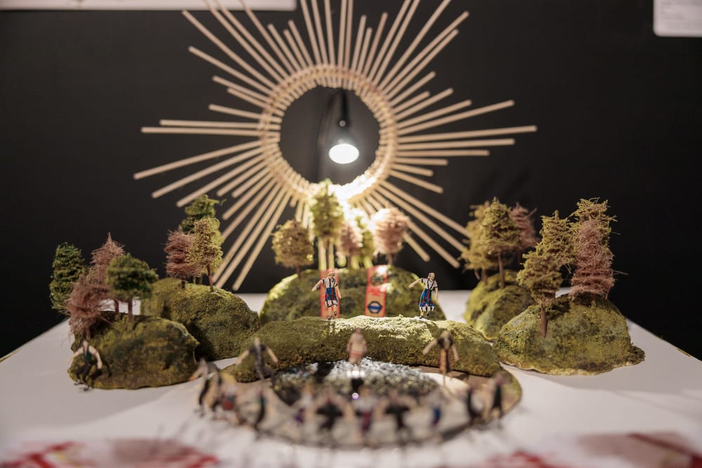 Close up photo of a set model showing six small hills dotted with trees. Two miniature people in Germanic traditional dress stand on the hill In the centre. At the back is a large representation of the sun from which an electric light illuminates the scene.