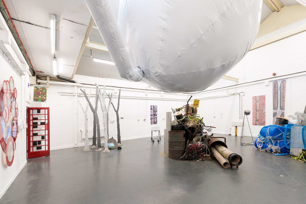 A large white room with a grey floor in which various works of sculpture are installed. To the left, a door in the style of the old British telephone boxes opens into another room. The outlines of tree trunks are cut from polystyrene. In the centre at the top of the image is a very large white inflatable sphere. Below this, a collection of found materials including wood and rolled up carpet are stacked in a pile.