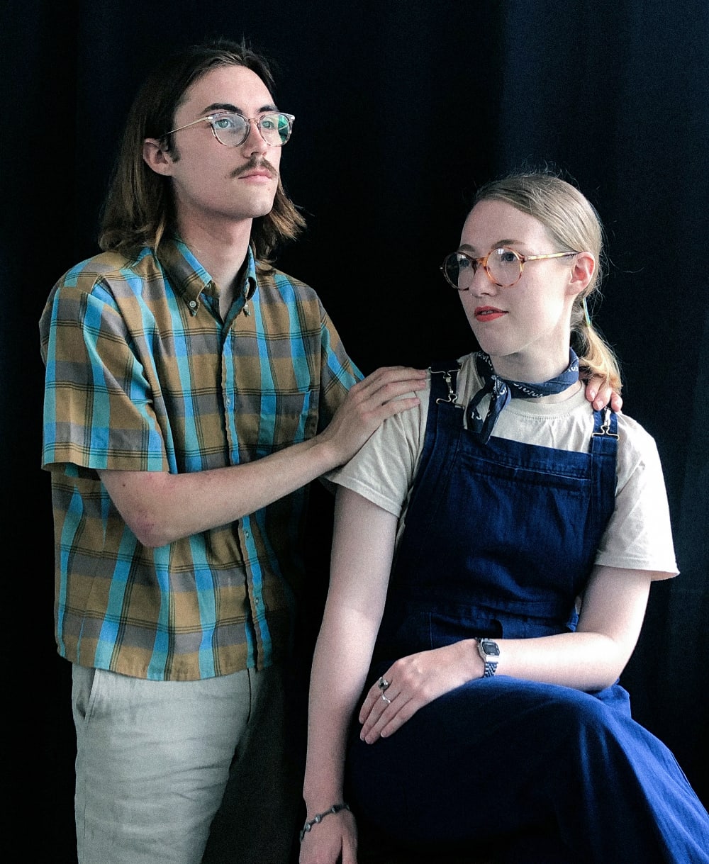 a couple pose for a photograph in a studio, he has his hands on her shoulders