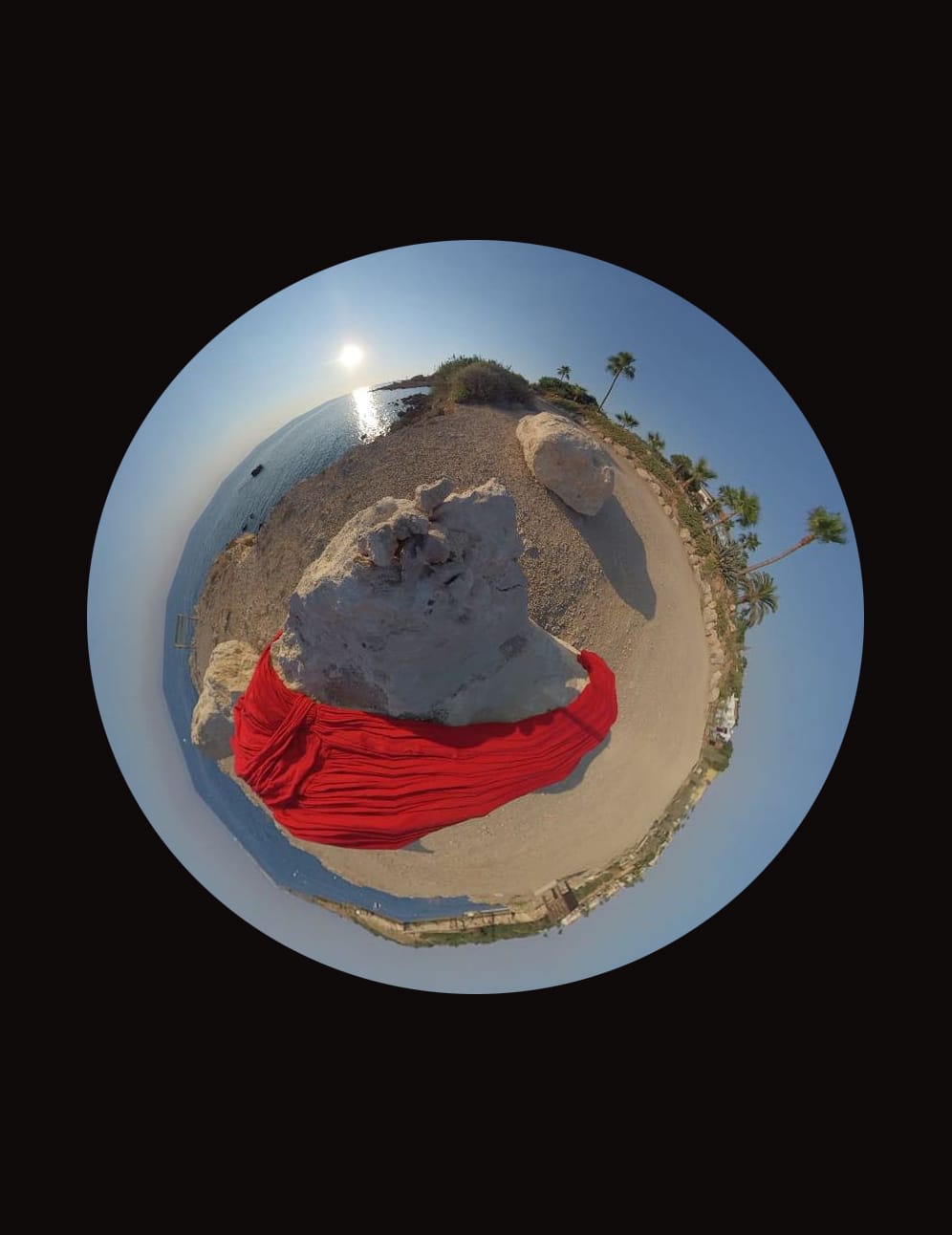 A spherical image of red fabric warps around a rock, in a seaside landscape.