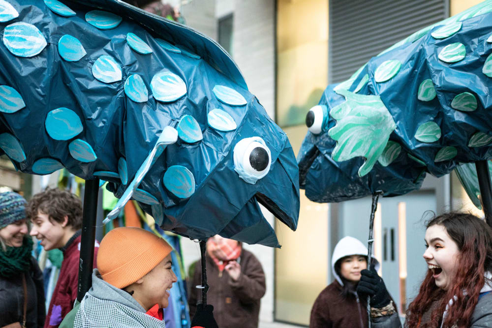 Two large blue spotted fish puppets made from paper and plastic are held above the heads of smiling students.