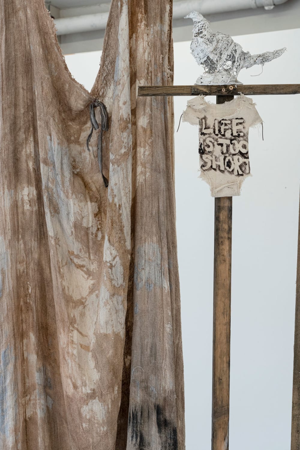 A painted piece of material hangs to the left of the frame. On the right, at the top of a wooden pole, is a baby grown painted with the words 'lifes too short' and a white wire bird sculpture.