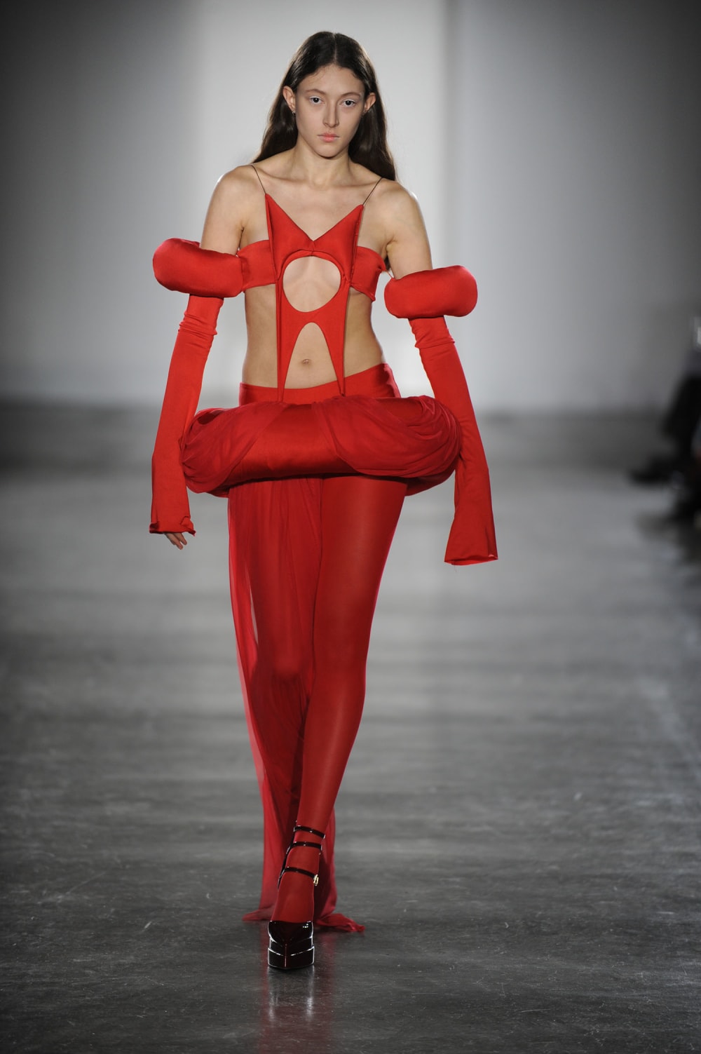 Woman on runway wearing red dress with big funky cut outs on bodice and off shoulder long sleeves and red tights paired with black heels