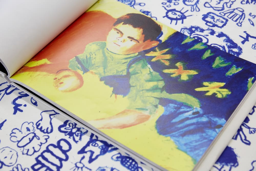 Image shows a colourful painting of small child holding an apple with flowers in the background. The child is wearing dungarees and a green floral top.