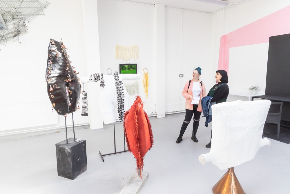 A white room in which sculpture is installed. The back wall is painting pink. To the right, two women stand enjoying four sculptures in black, white, red and white.