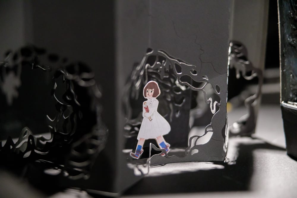 Detail shot of a set model showing dark grey card with holes cut in it in the style of tree roots. In the middle of the image is an illustration of a small girl in a white dress, blue socks and brown shoes. She has a grumpy expression and is clutching a doll in her crossed arms.