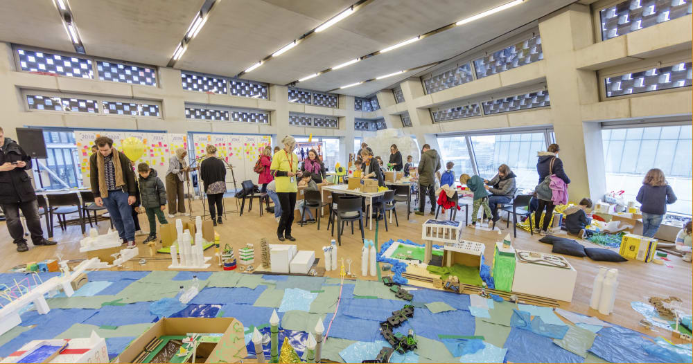 A room full of people standing and sitting at tables and chairs scattered throughout. In the foreground is a map made from waste materials laid out on the floor, spanning the whole room.