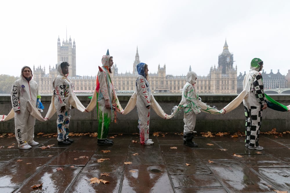 A line of people wearing boilersuits which are all joined together standing on the south bank of the Thames with the Houses of Parliament visible in the background.