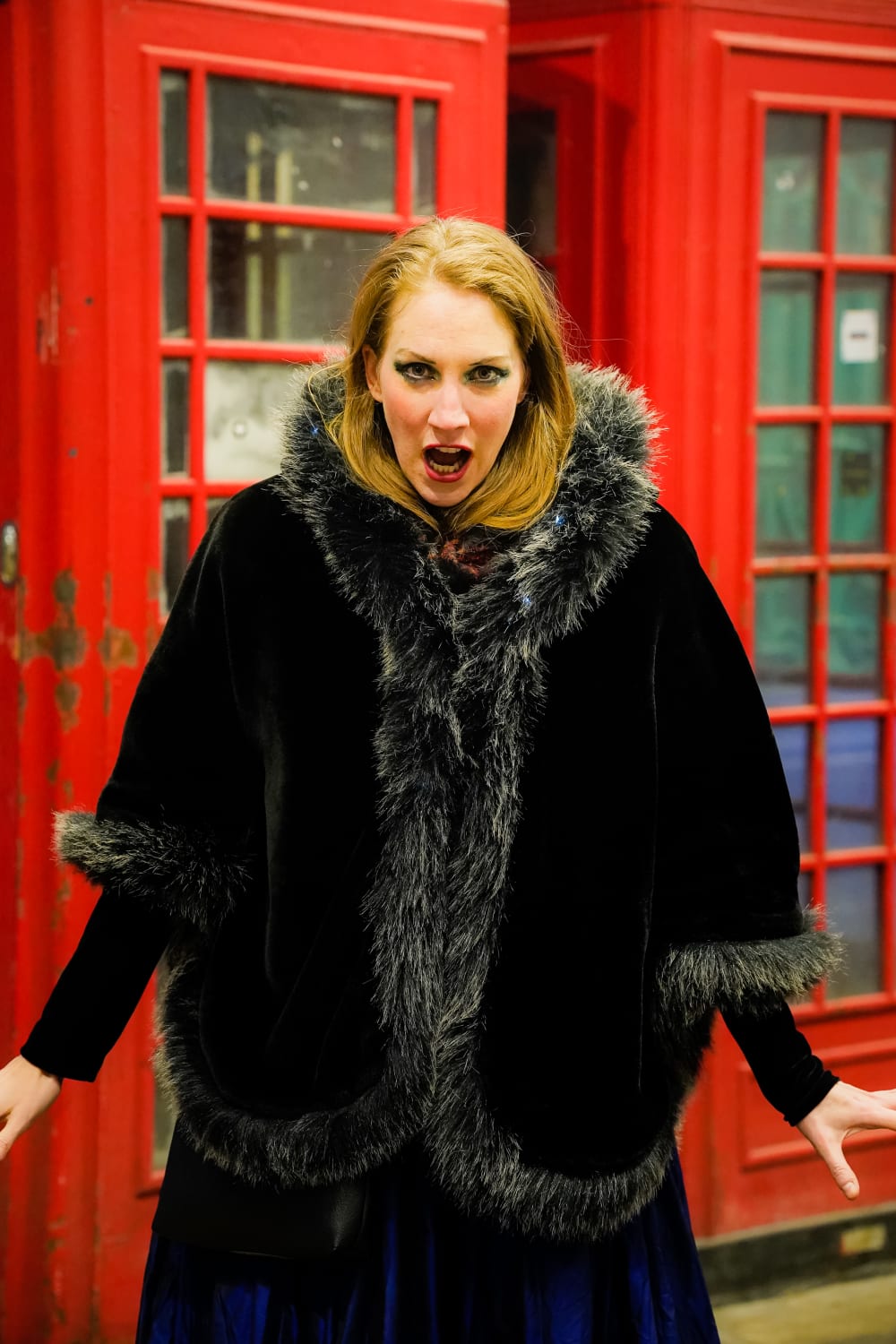 Maria in a long black coat, looking into the camera. She's stood in front of two red phone boxes