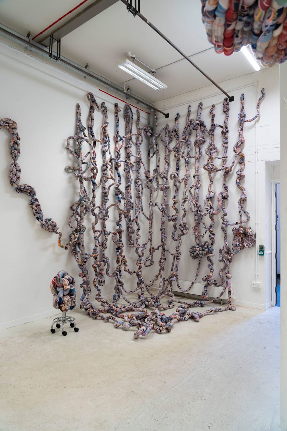 An artwork hanging the full height of two adjacent walls in a gallery space. The work looks to be made of fabric ropes curling and hanging in tendrils down the wall.