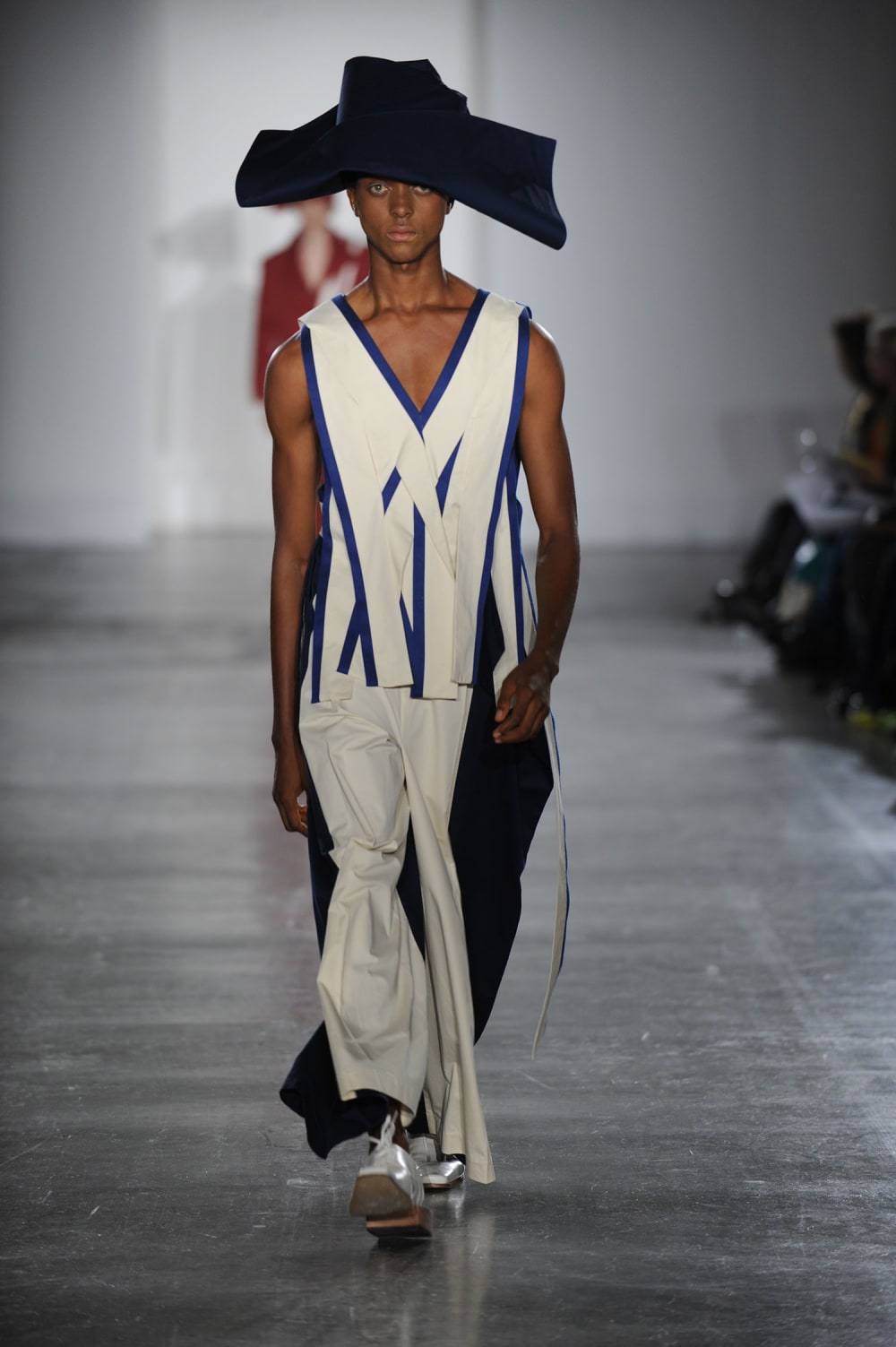Man on runway wearing nay blue draped hat, white vest top with blue detailing criss cross panels paired with navy blue pants with white overlay detail and silver boots