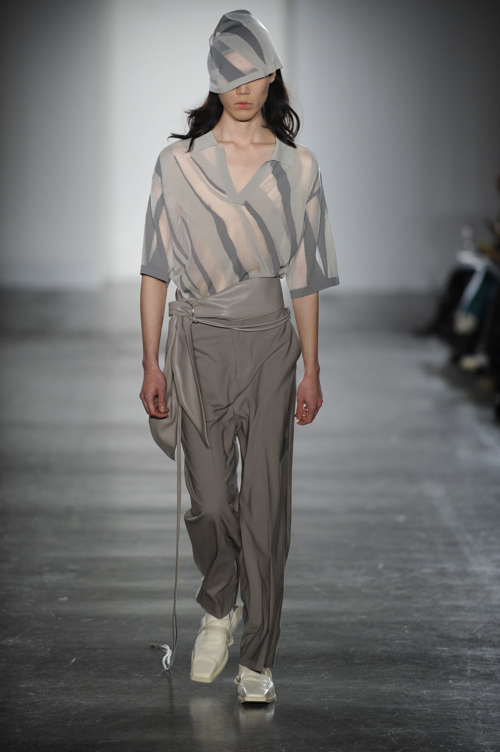 Man on runway wearing see through stripped shades of grey Juliet cap style hat, matching material open collared elbow length t-shirt, grey faux leather belt tied to the side with silver hoop buckle, dark warm grey straight cut pants and white shoes