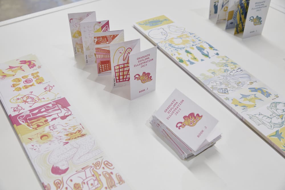 Image shows a close-up of a table with 2 long pieces of illustrated paper lying on either side of the shot. In the middle is a tiny fold-out booklet which reads ‘Graduate Diploma Illustration 2023’