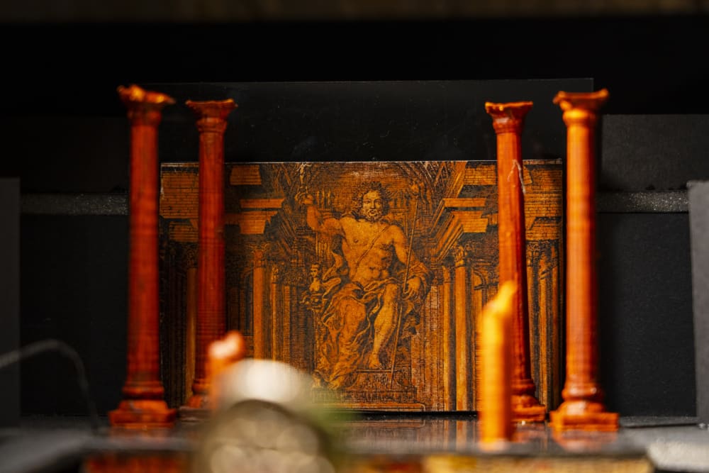 A miniature model installation of red pillars in front of a painting of a shirtless man sitting on a throne