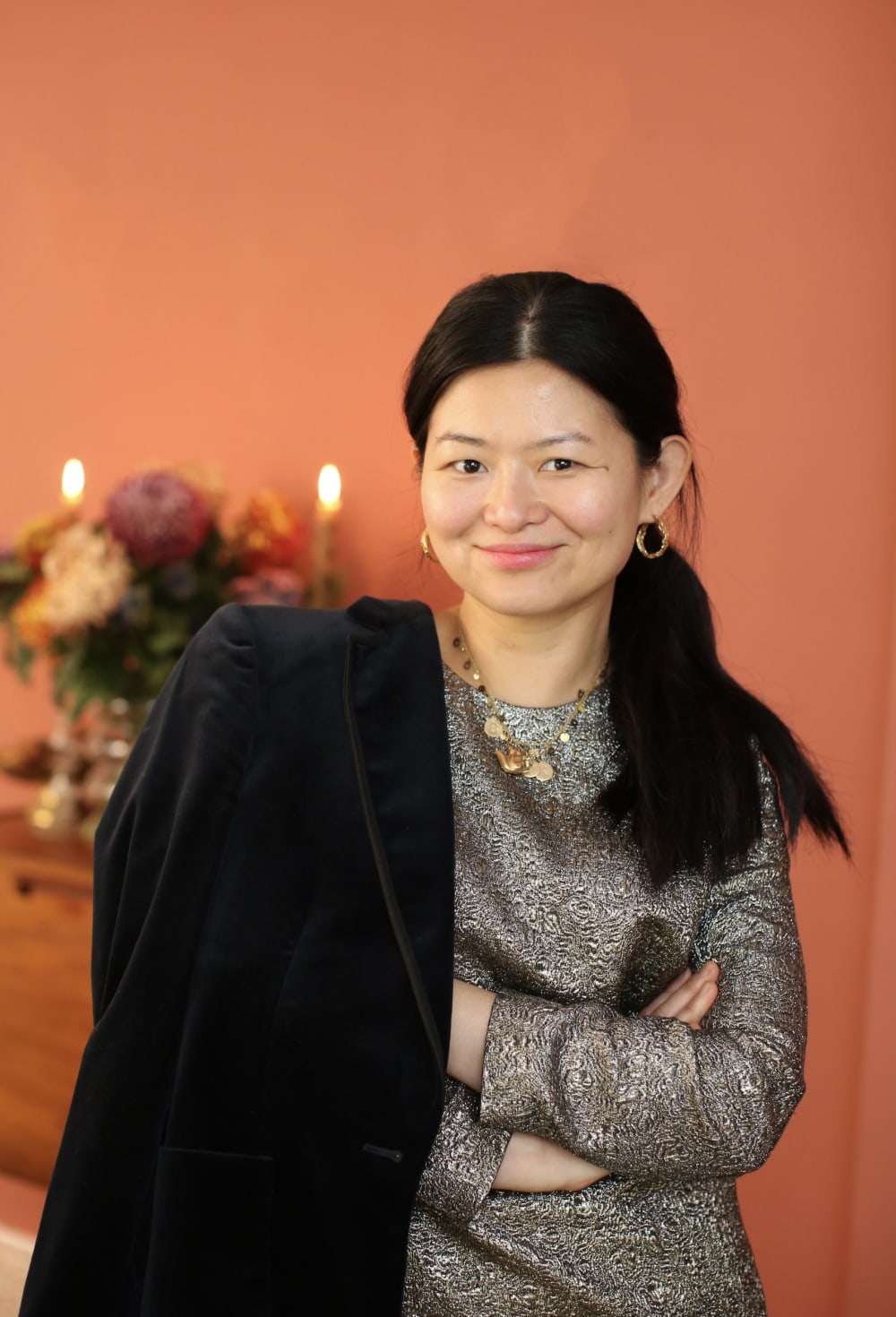 Kuo-Fang wearing a dark metallic top, with a black coat over her right shoulder. She's got her arms crossed. 